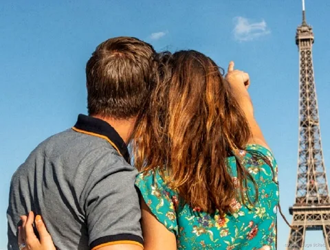 Couple seen from behind pointing up at the Eiffel Tower under a blue sky, part of a day with Moulin Rouge tickets