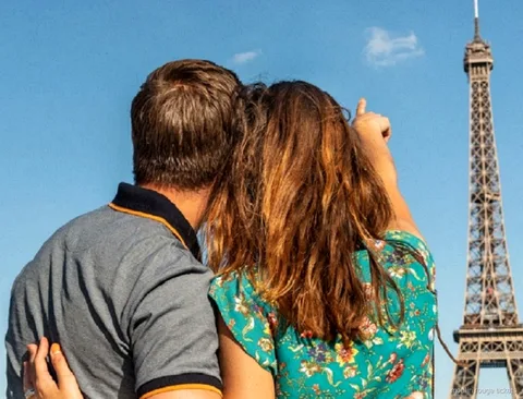 Casal de costas apontando para a Torre Eiffel sob céu azul, parte do dia com bilhetes Moulin Rouge em Paris