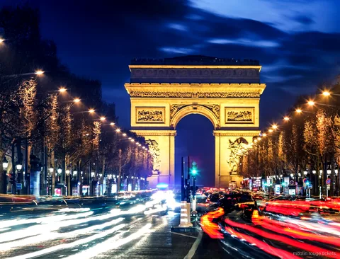 Triumfbågen upplyst på natten vid slutet av Champs Élysées med ljusspår från trafiken, rutten före Moulin Rouge-biljetter