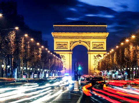 Arco di Trionfo illuminato di notte in fondo agli Champs Élysées con scie luminose, tappa prima dei biglietti Moulin Rouge